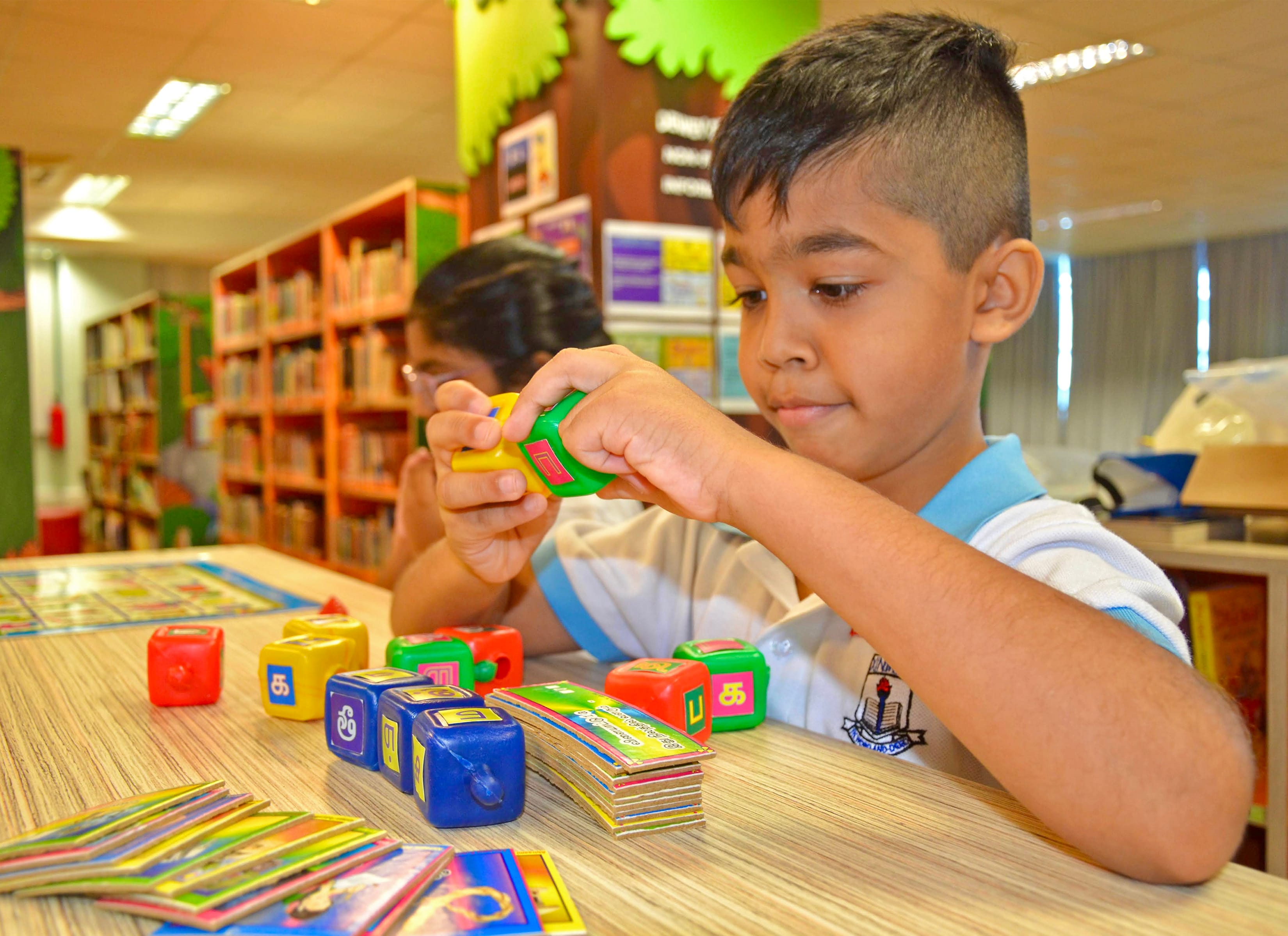 A young boy plays with colorful letter blocks and cards at a table in a library with bookshelves behind him.